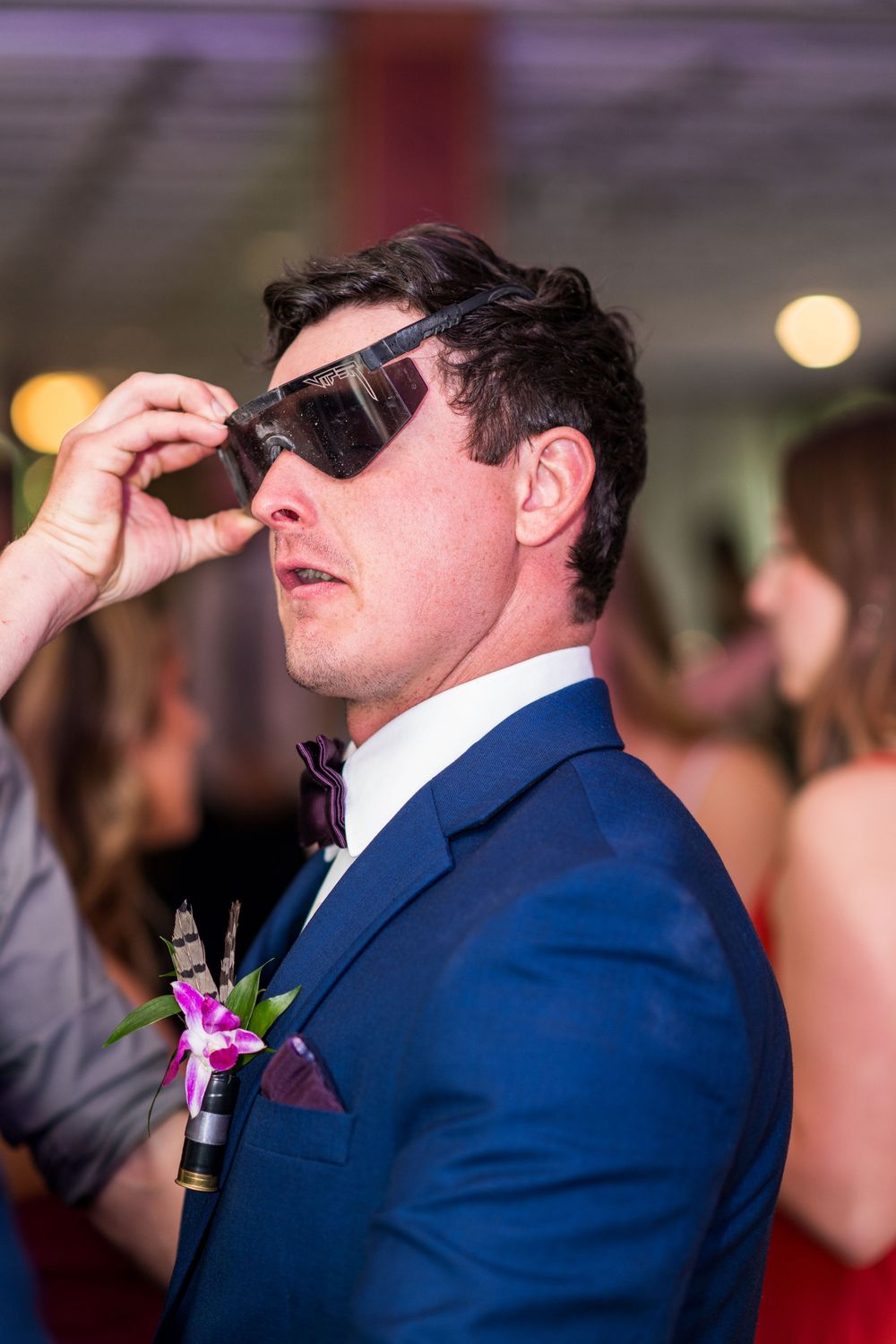 Stylish groom in blue suit with floral boutonniere adjusting sunglasses at wedding reception.