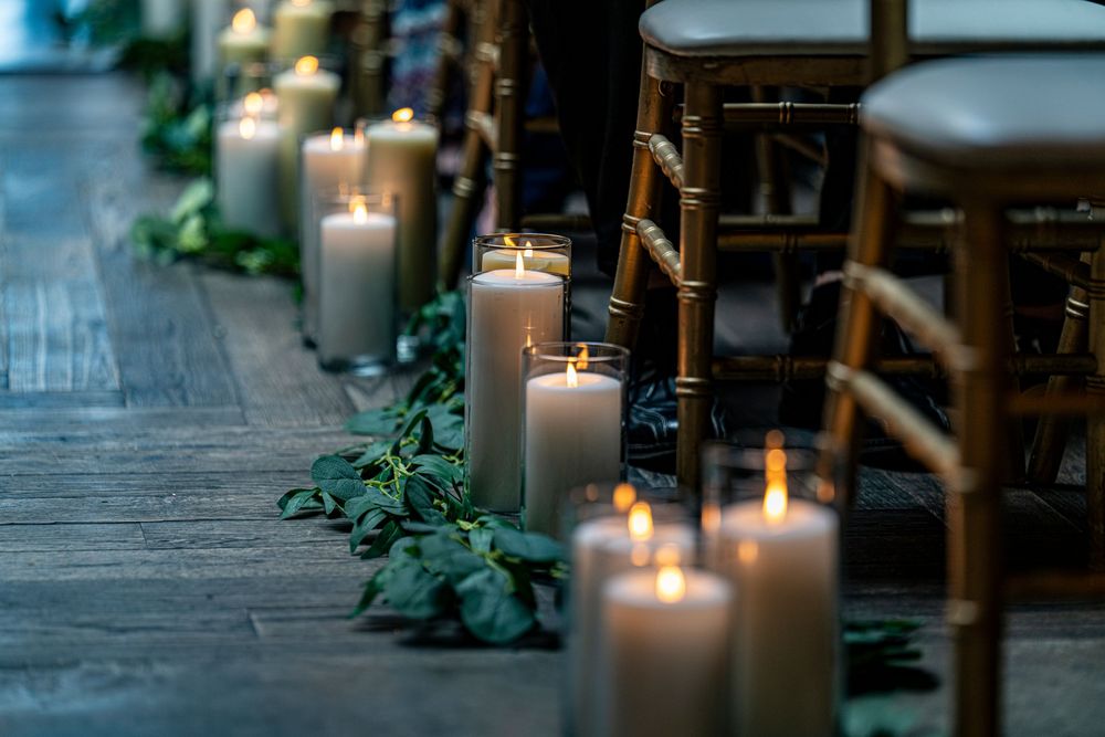 Elegant wedding aisle lined with glowing white pillar candles and greenery beside wooden Chiavari chairs.