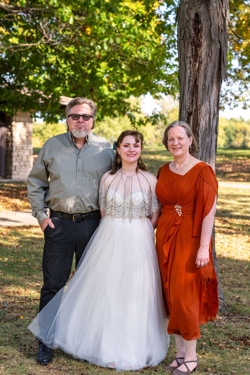 Bride in white gown poses with parents outdoors near a tree during a wedding celebration.