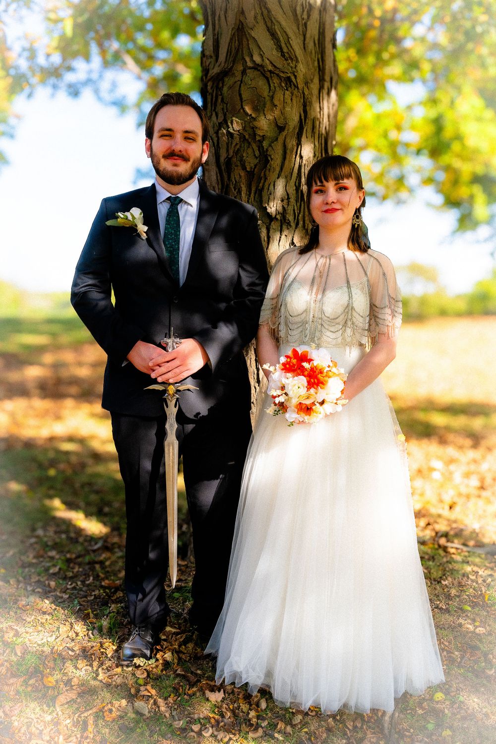 Bride and groom standing by large tree trunk in autumn park during outdoor wedding portrait.