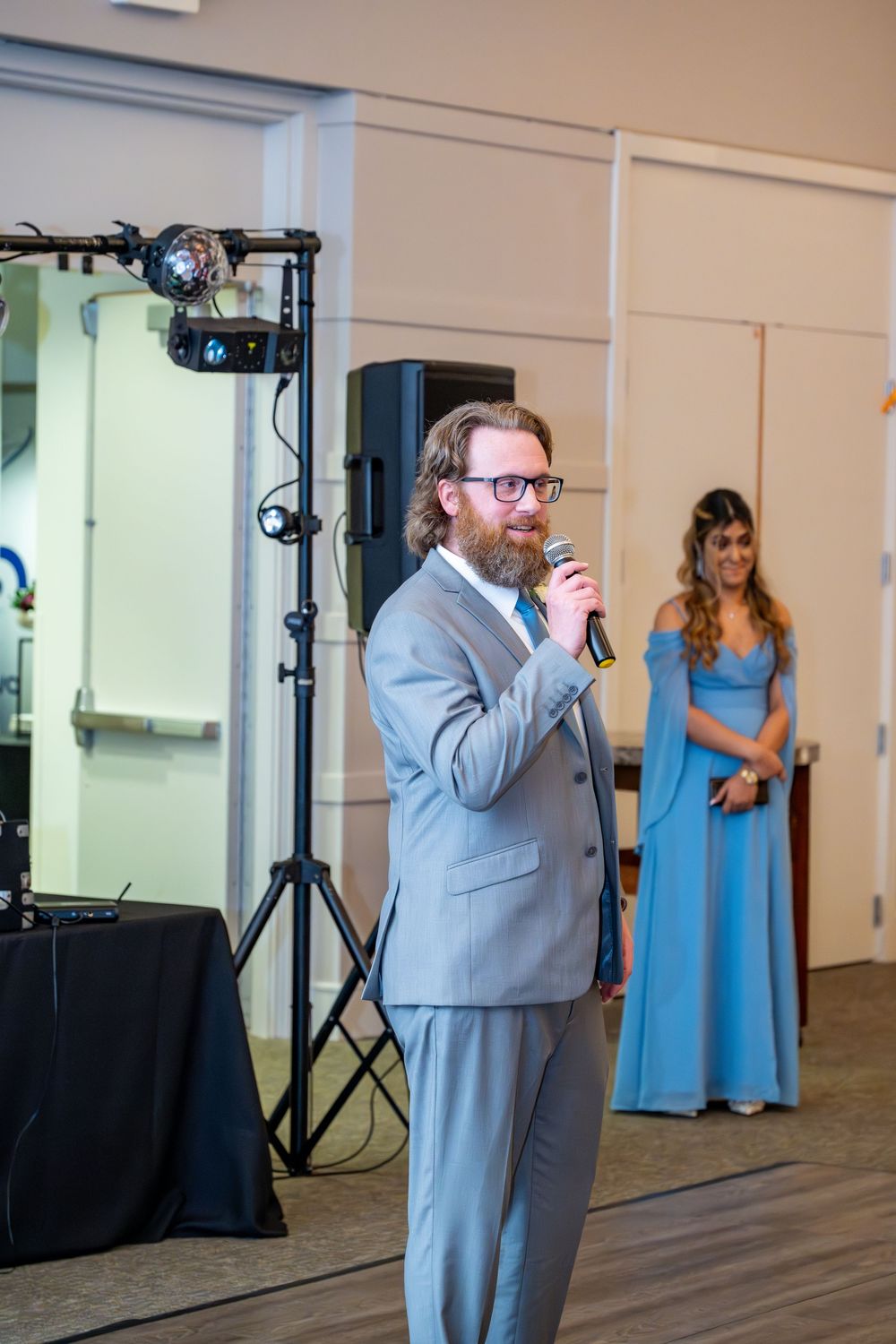 Bearded man in gray suit giving speech at wedding reception with microphone, woman in blue gown visible in background.