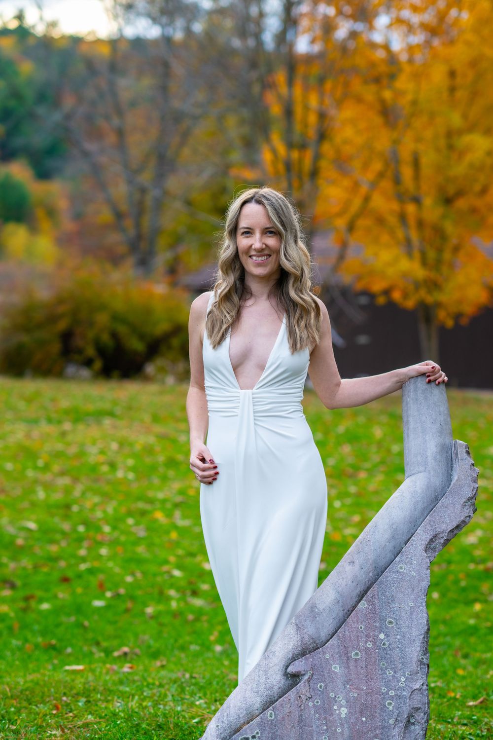 Smiling bride in a white deep-V gown posing outdoors with vibrant autumn foliage in the background.