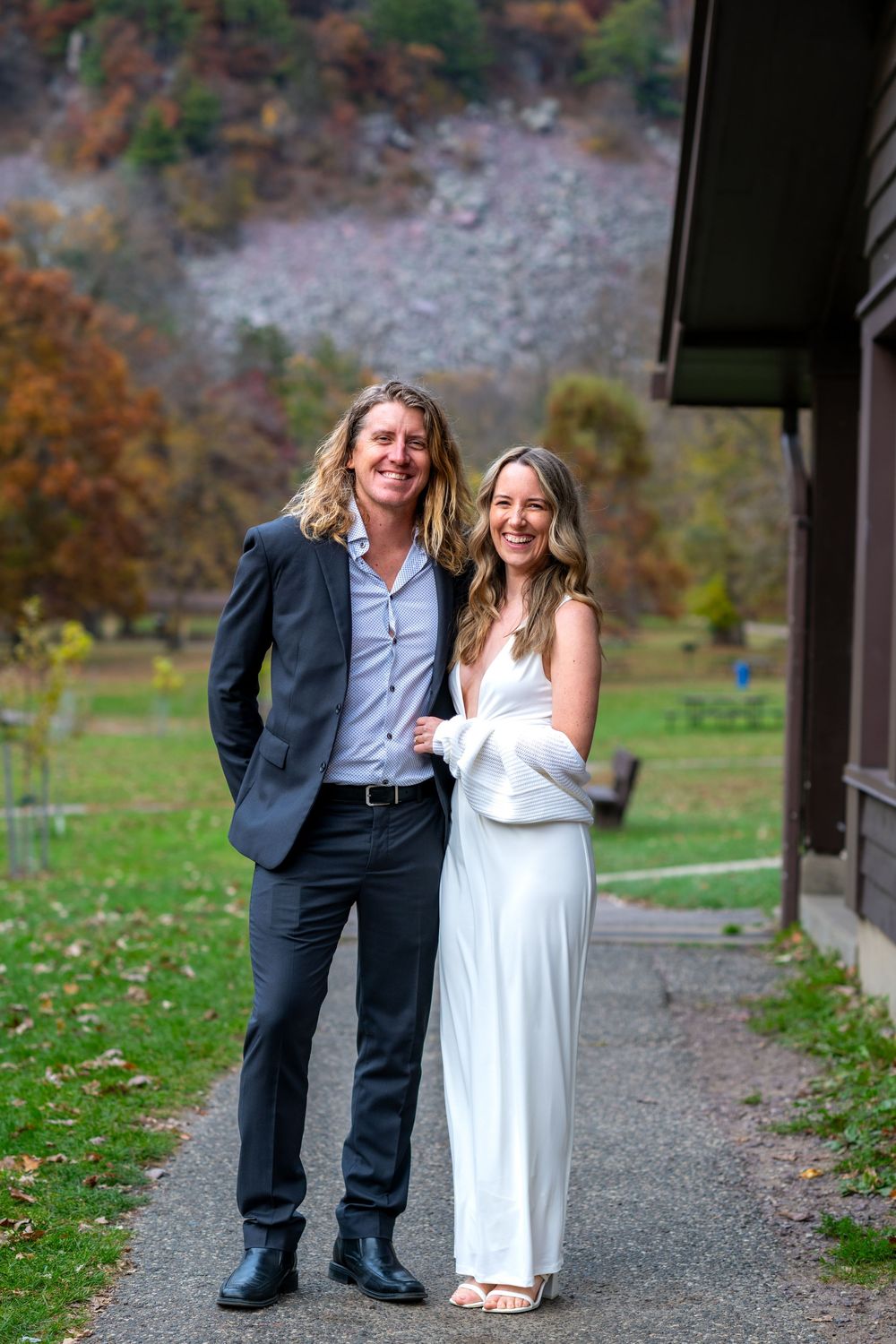 Smiling bride and groom posing together outdoors near a rustic building with autumn mountain backdrop.