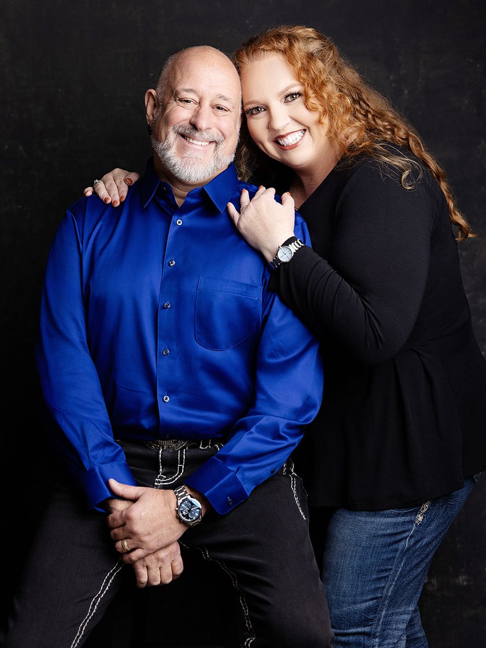 A couple in a studio portrait setting against a black background, with one wearing a bright blue shirt.