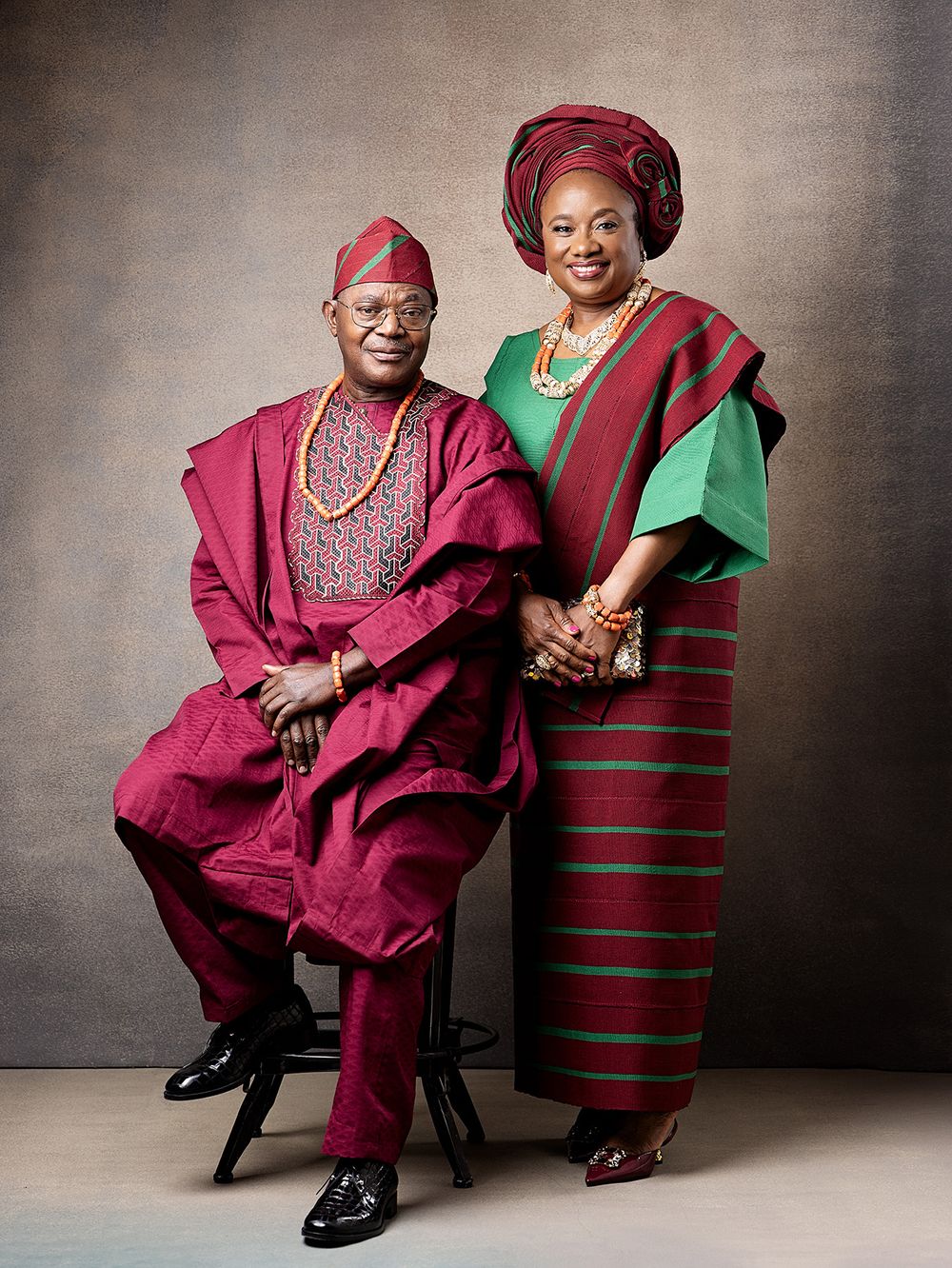 A couple in traditional Nigerian attire poses in burgundy and green formal wear with cultural headpieces and jewelry.
