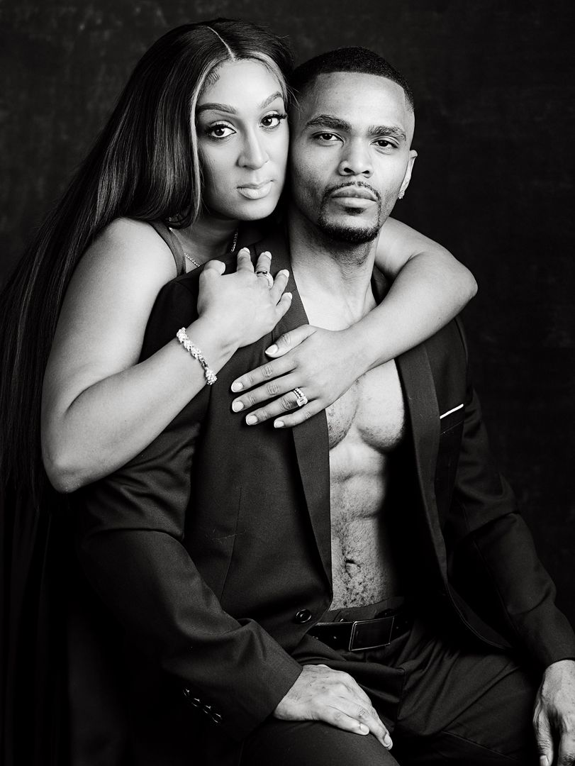 Black and white studio portrait of an elegant couple, woman in black dress embracing a shirtless man in suit jacket.