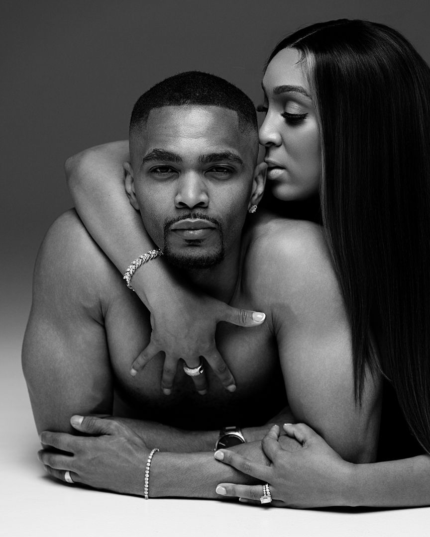 Black and white portrait of a shirtless man wearing jewelry with a woman embracing him from behind in a studio.