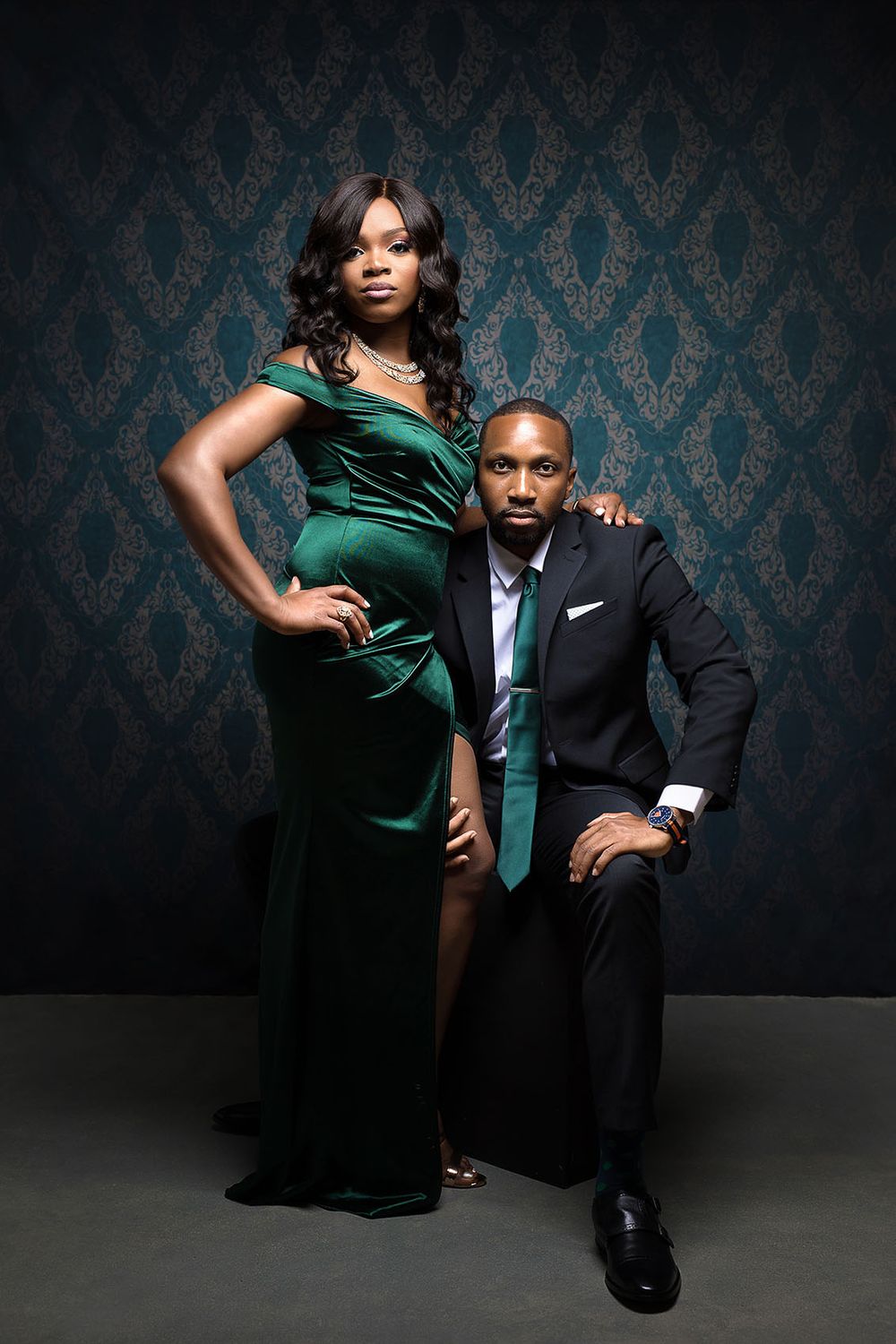 Stylish couple poses together against dark damask wallpaper in elegant emerald dress and black suit.