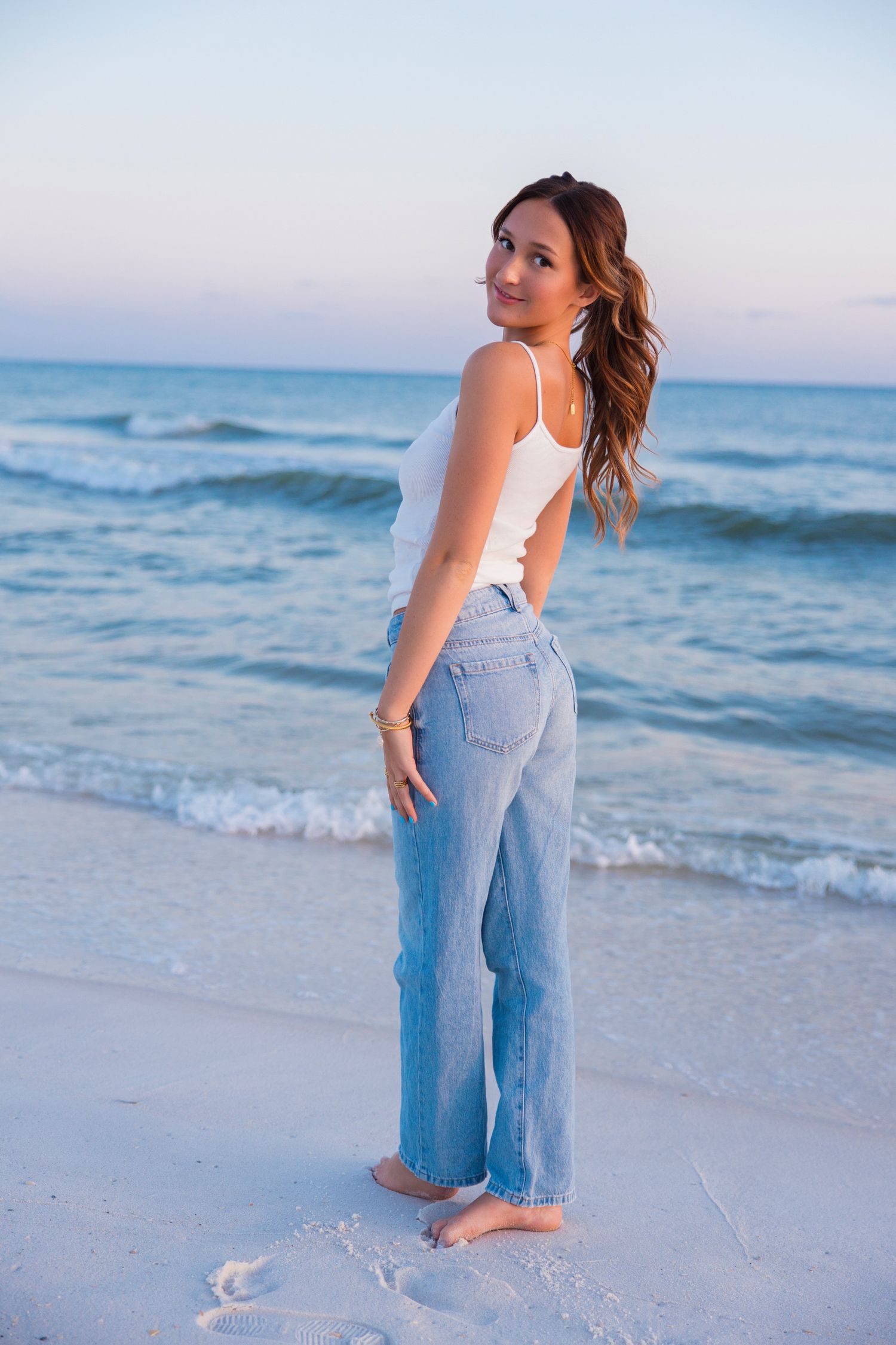 Person wearing denim overalls stands at the beach during sunset while waves crash on the shore.