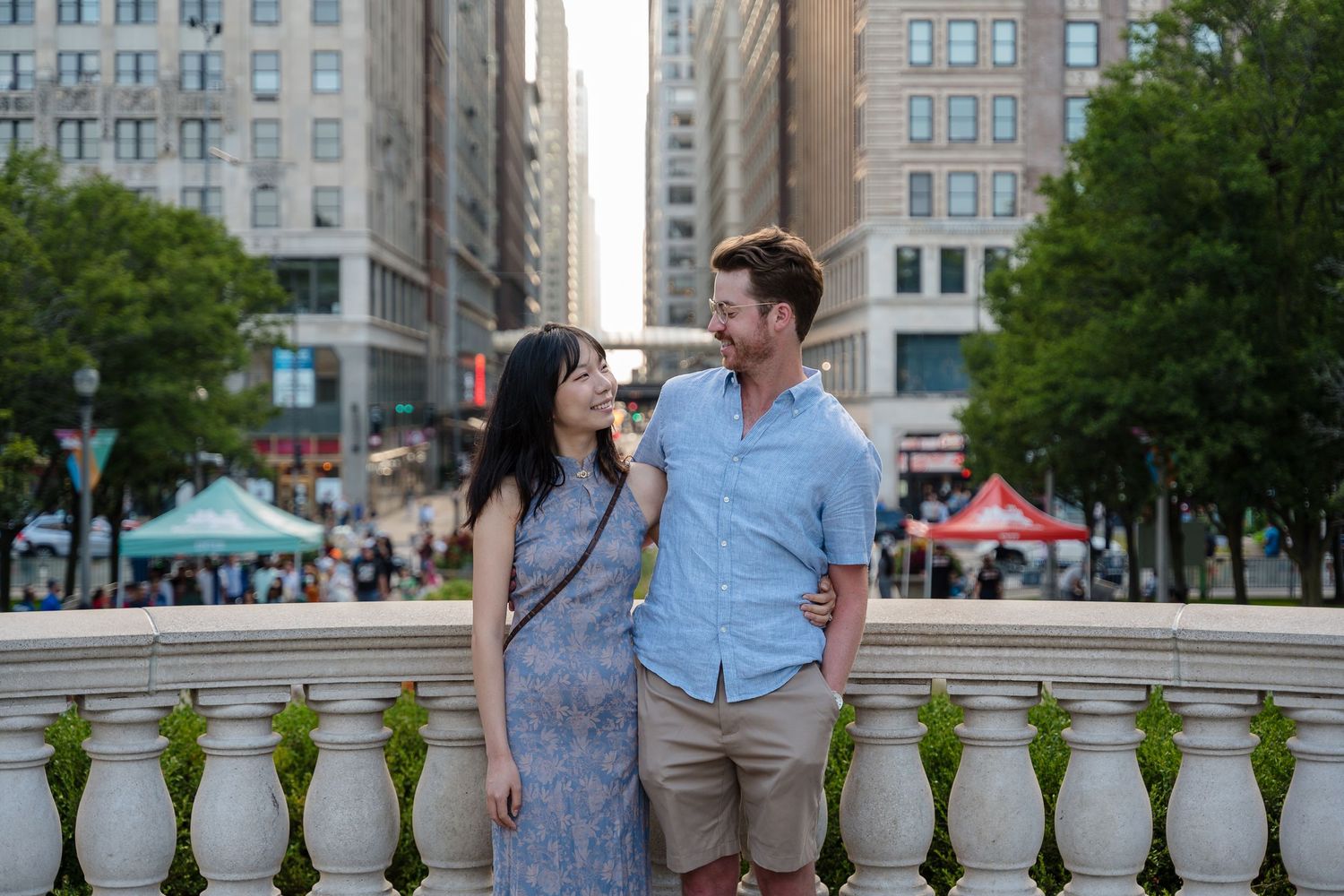 Couple strolling along a city sidewalk with tall buildings and red umbrellas in the background.
