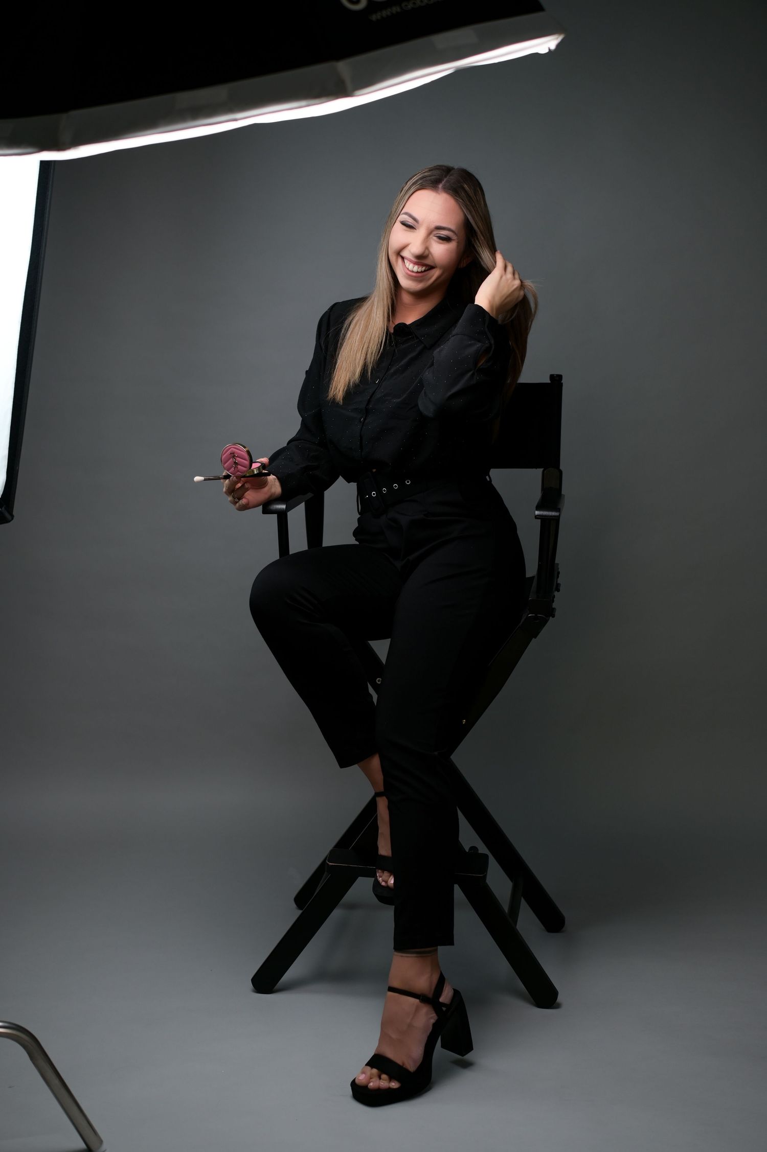 A person in all black clothing poses playfully on a director's chair against a gray studio backdrop.