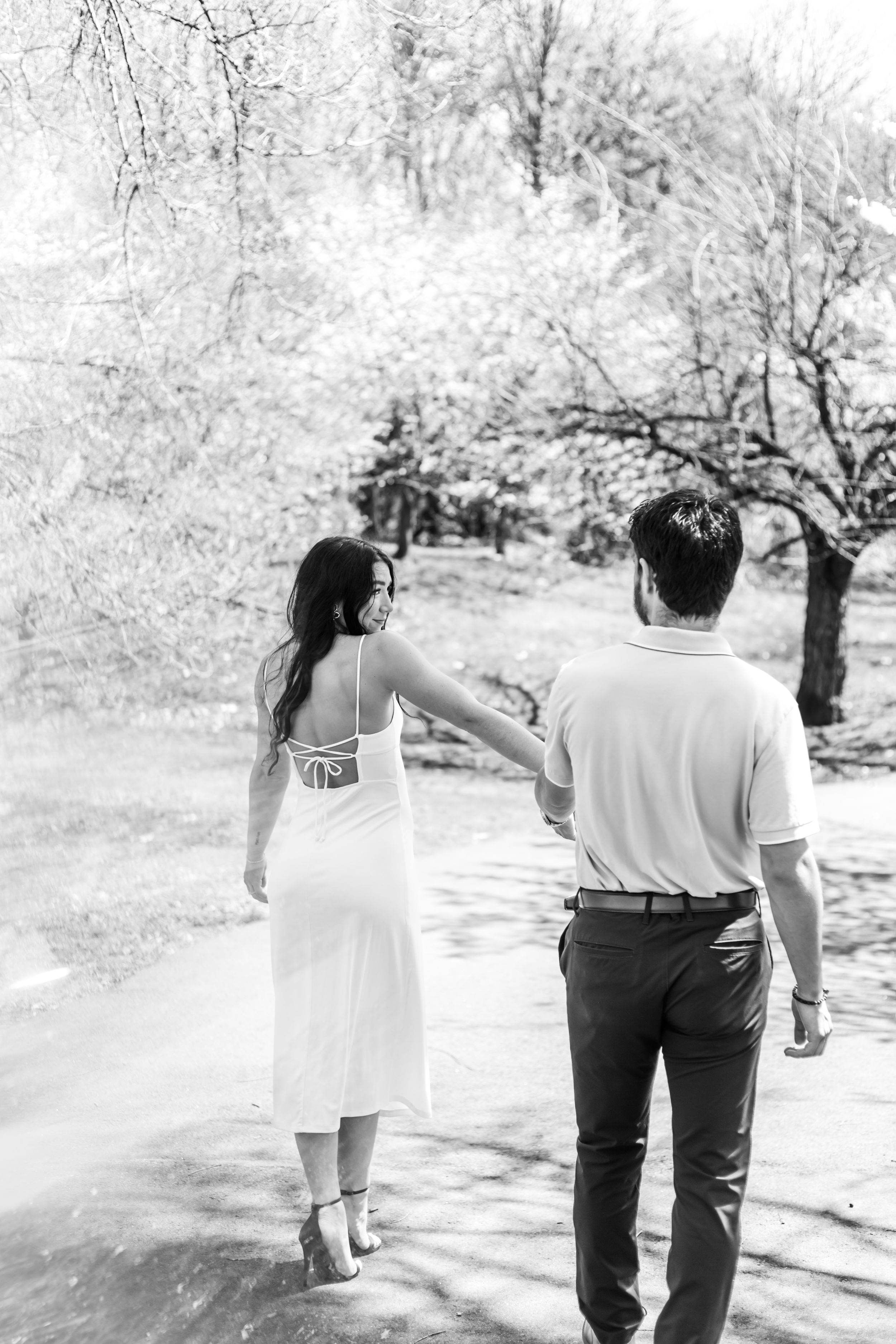 A couple walks hand in hand along a sunlit path beneath flowering spring trees.