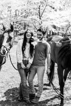 A couple shares joyful moments together during an outdoor photoshoot with a black horse in a natural setting.