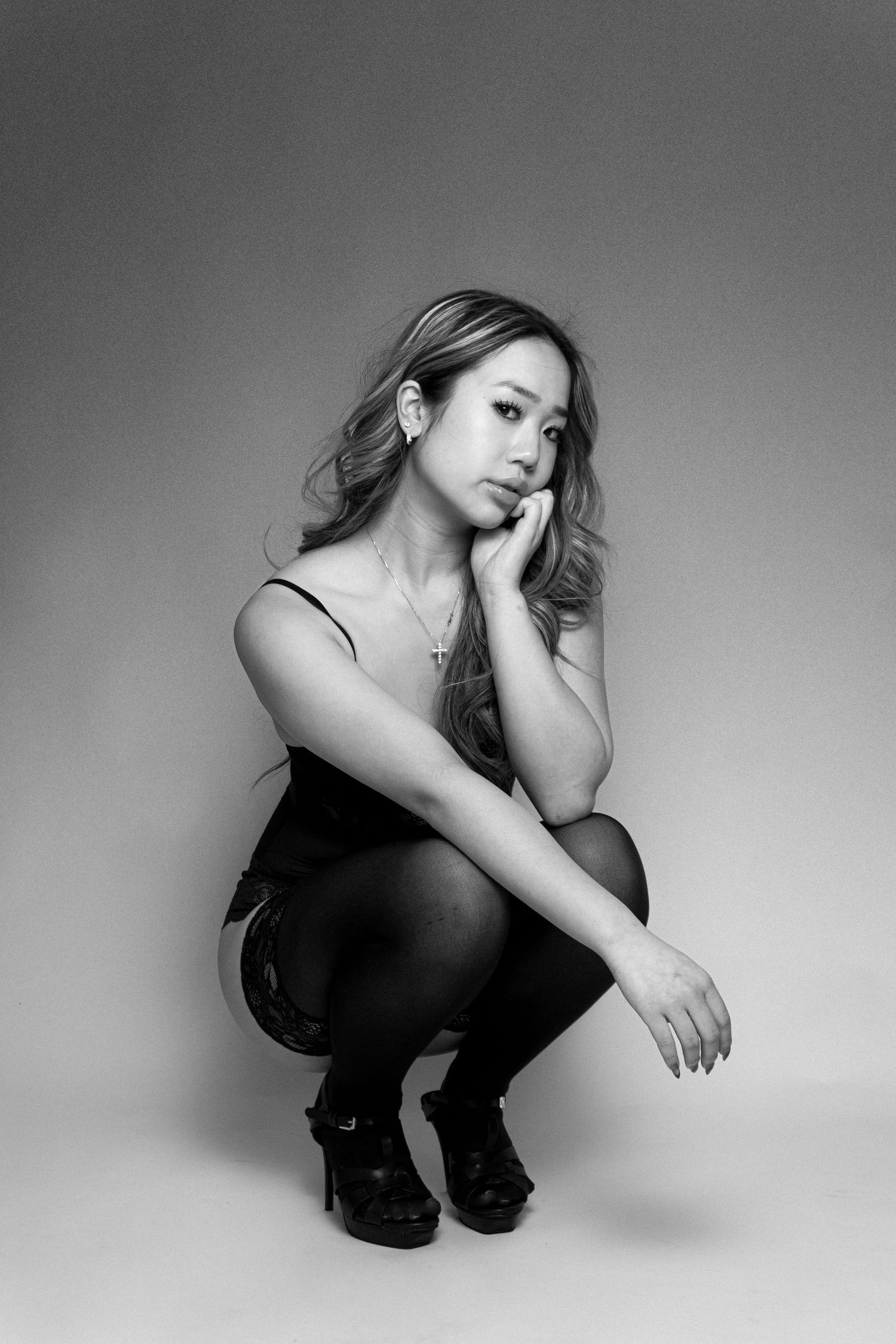 Black and white studio portrait of a young woman crouching in heels and a black dress, resting chin on hand.