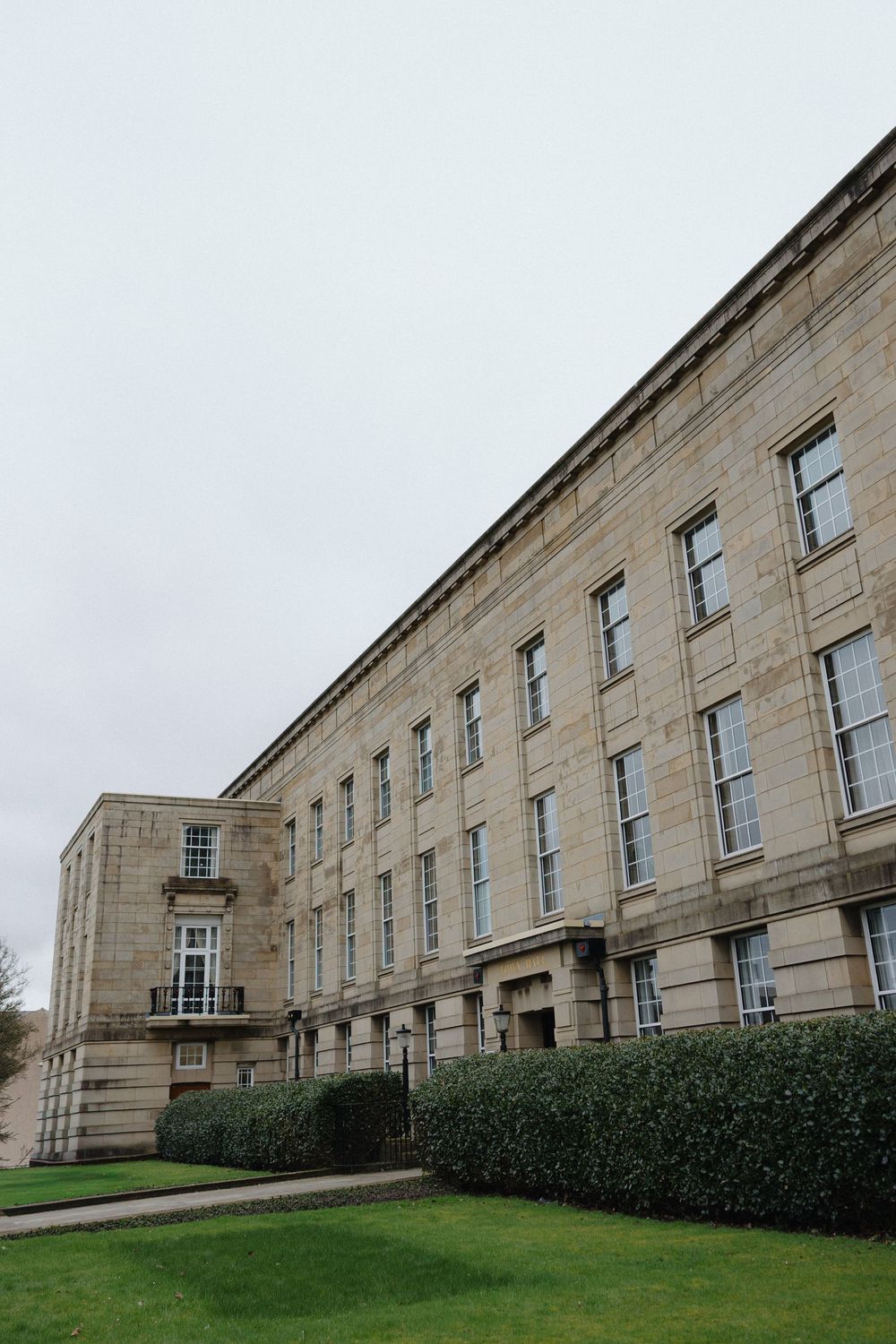 Stone building with Georgian architecture features rows of symmetrical windows and a sloping lawn on a cloudy day.