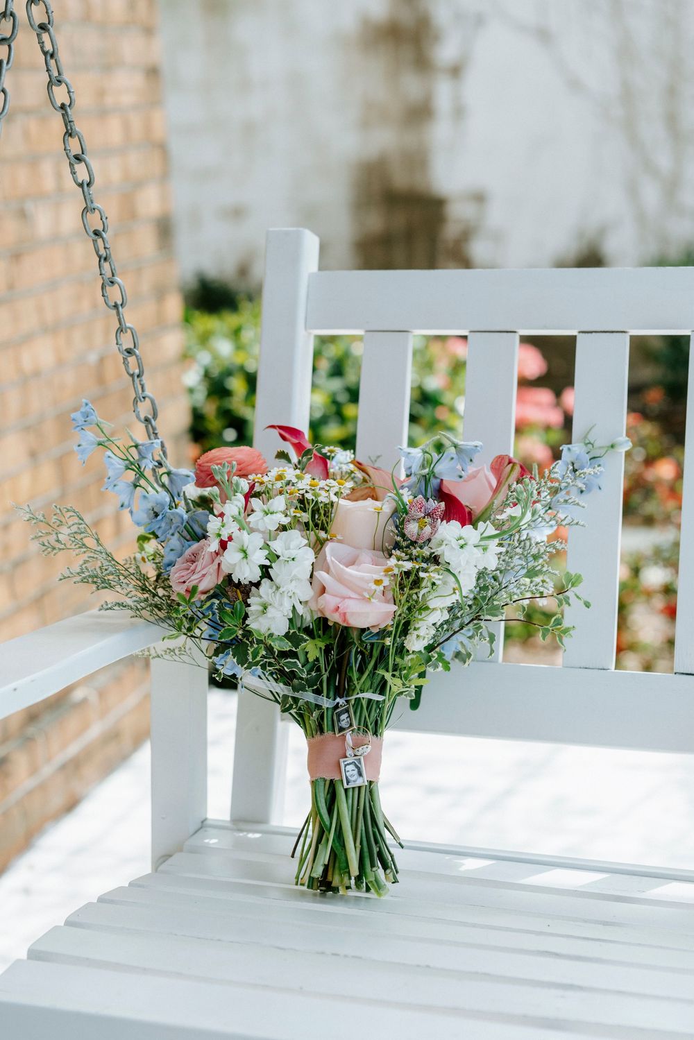 A delicate bouquet of pink roses and white wildflowers sits on a white porch swing with a chain.