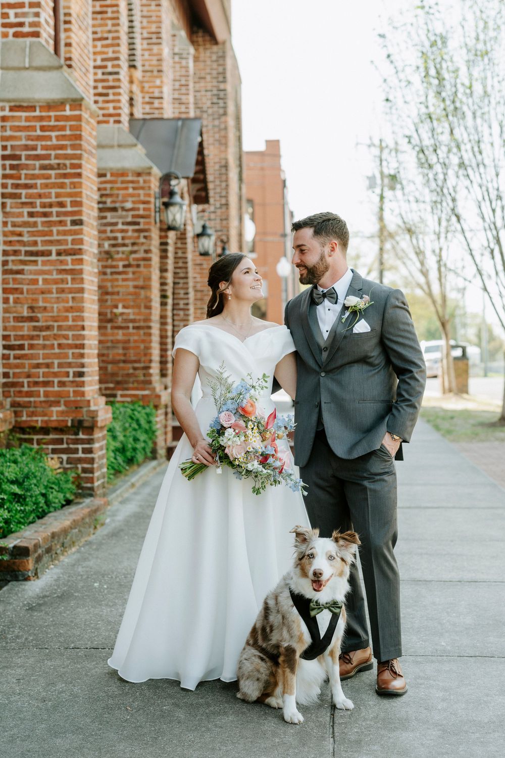 A couple in wedding attire poses outside a brick building with their Australian Shepherd dog wearing a bow tie.