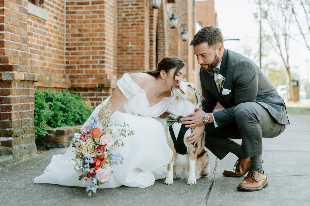 A couple in wedding attire kneels down to pet their white dog outside a brick building.