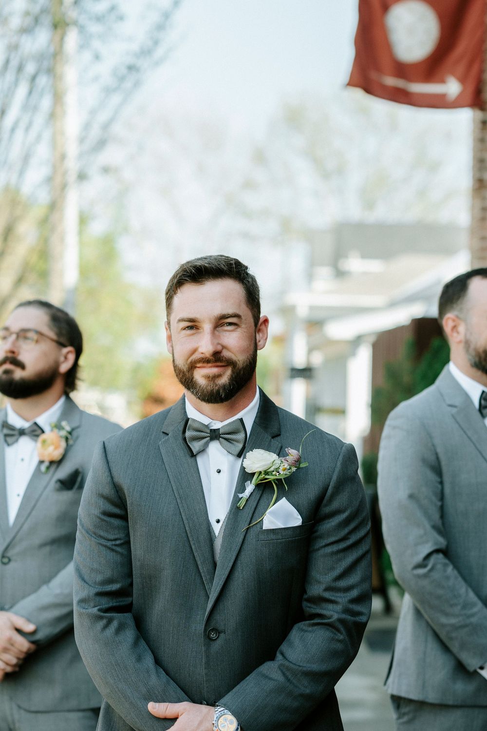 A groomsman in a charcoal gray suit with bowtie and boutonniere stands at attention during an outdoor wedding ceremony.