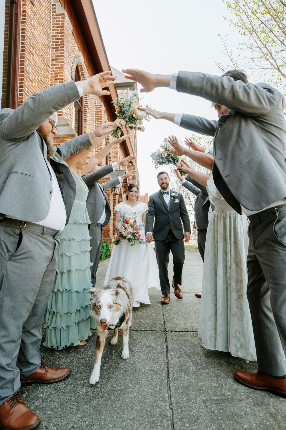 Wedding party in gray and sage green attire performing synchronized dance moves outside a brick building.