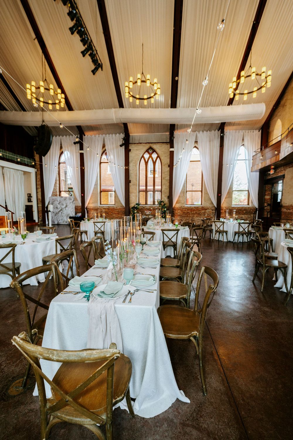 Elegant rustic wedding reception venue with white tablecloths, wooden cross-back chairs, and warm pendant lighting.