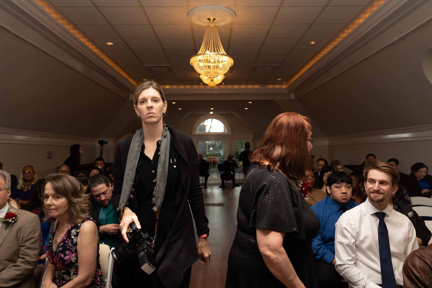People gather in an elegant ballroom with a crystal chandelier during what appears to be a formal event.
