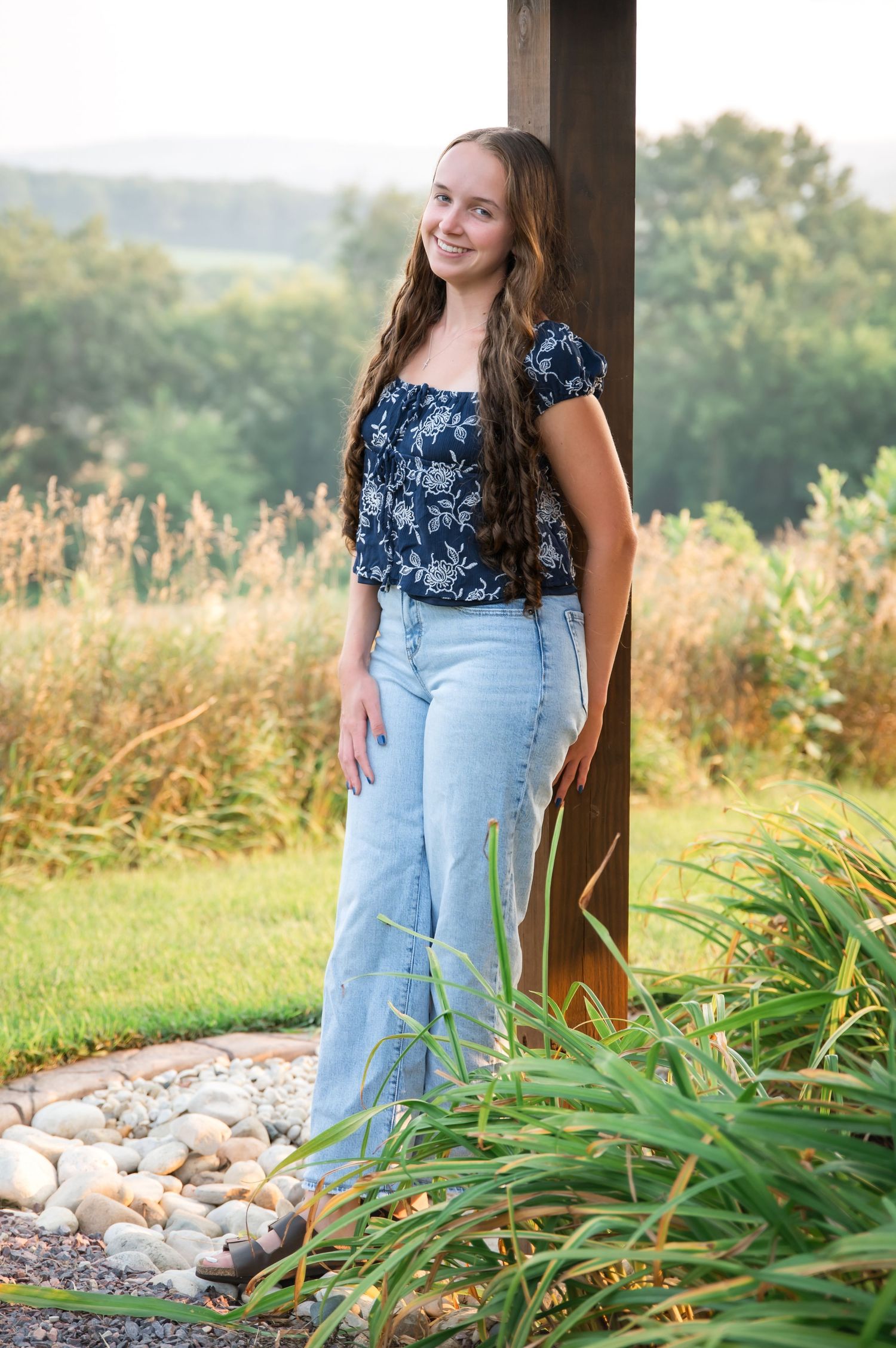 A person in a blue floral top and light denim jeans poses outdoors near tall grass and rocks at sunset.
