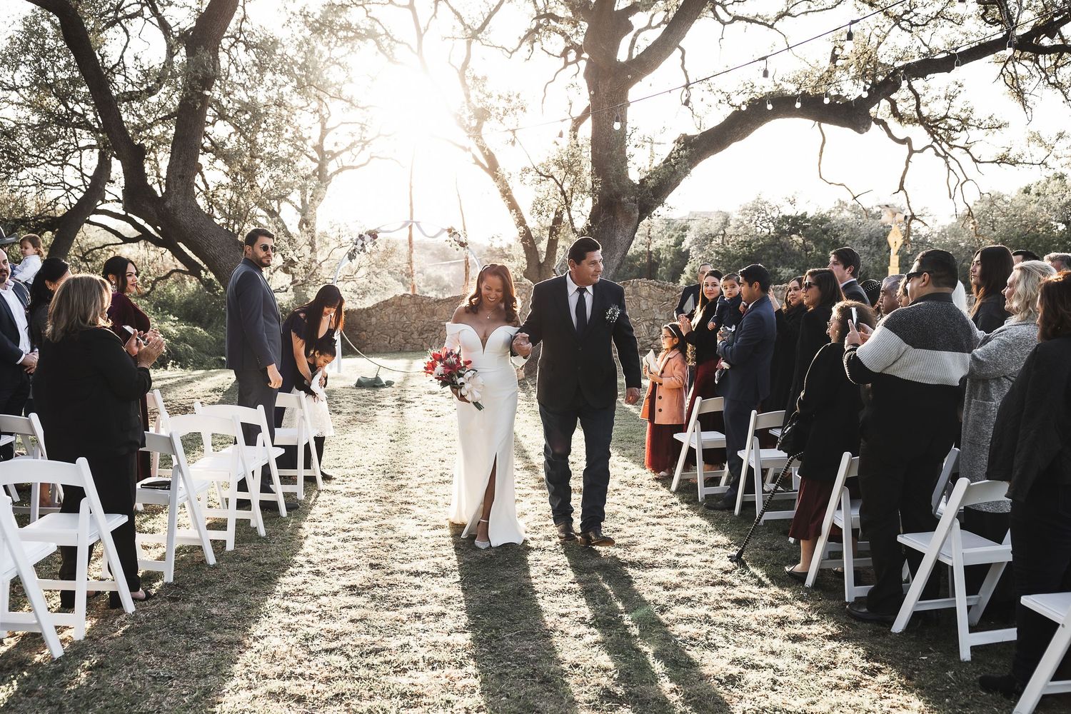 Guests gather under a sunlit tree for an outdoor wedding ceremony with white chairs arranged in rows.