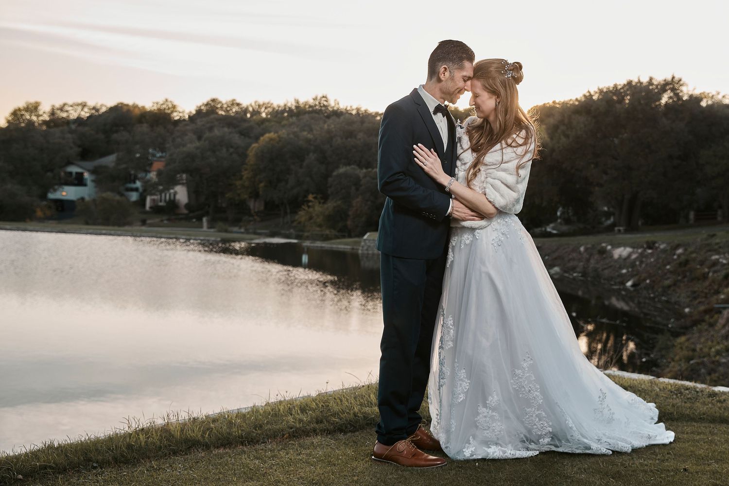 A couple embraces during their lakeside wedding at sunset, with the bride wearing a flowing white lace gown.