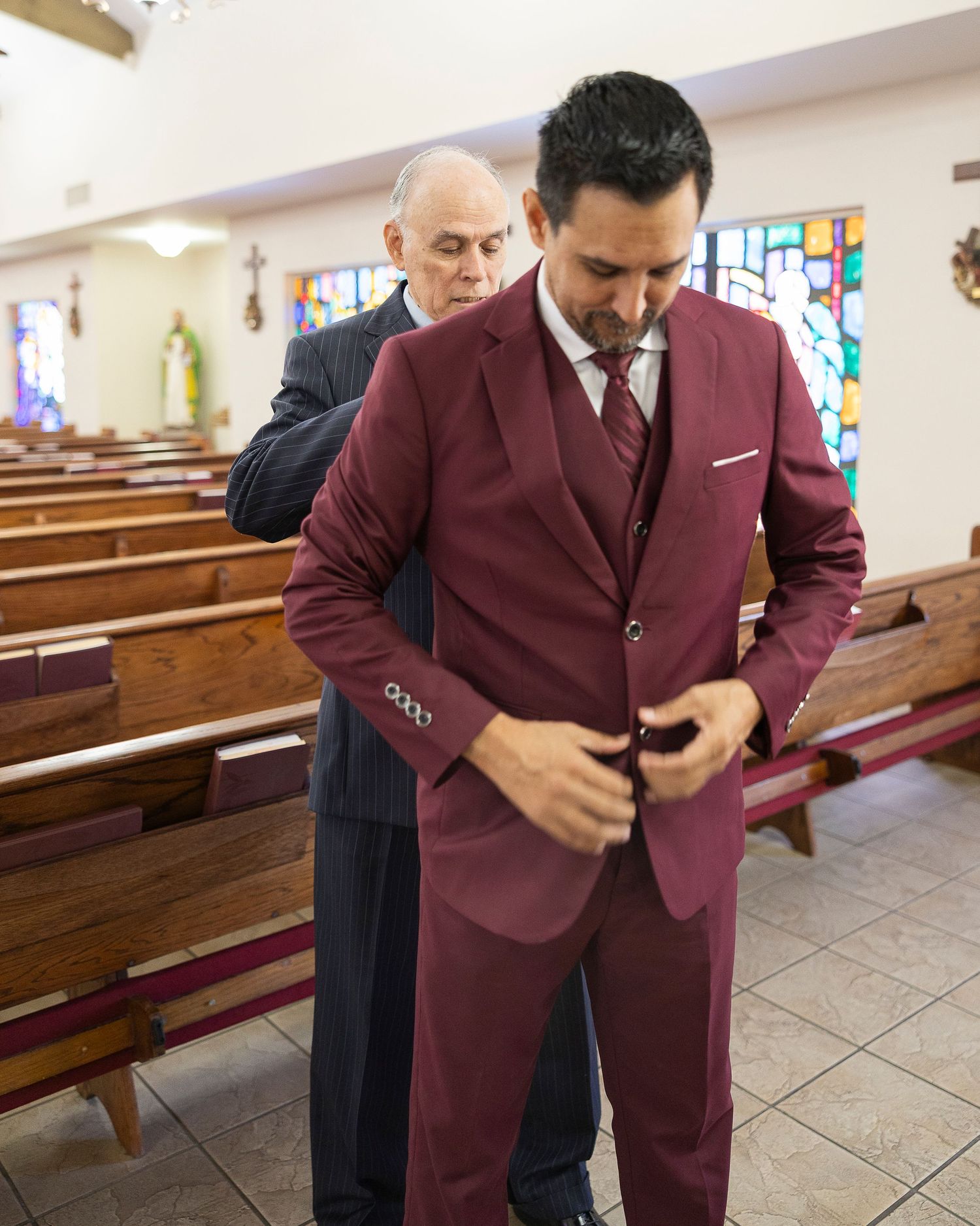 A person in a burgundy suit checks their phone while standing in a church with wooden pews and stained glass windows.
