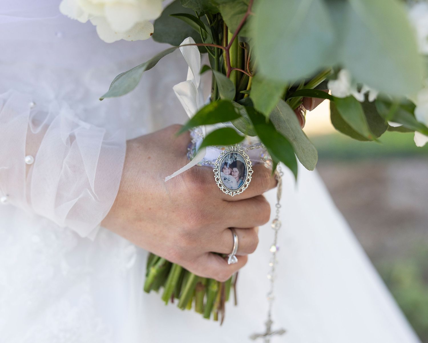 A white bridal bouquet held with hands wearing a large oval engagement ring and wedding band.