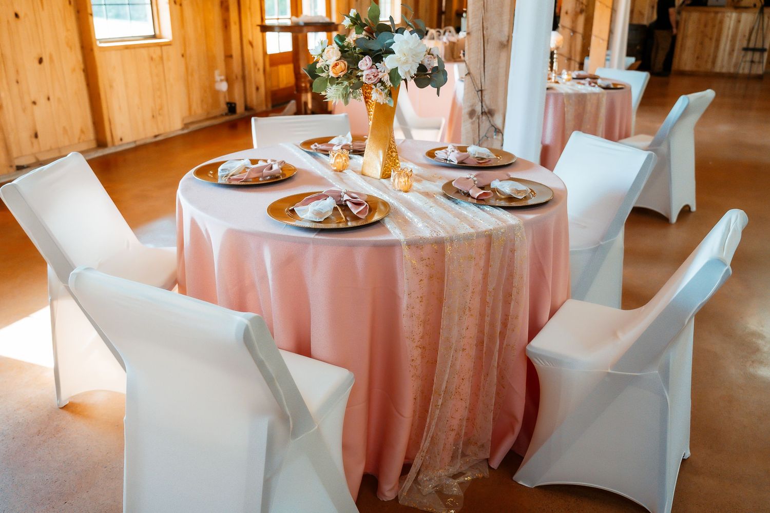 Elegant wedding reception setup in rustic wooden barn with white chair covers and soft pink tablecloths.