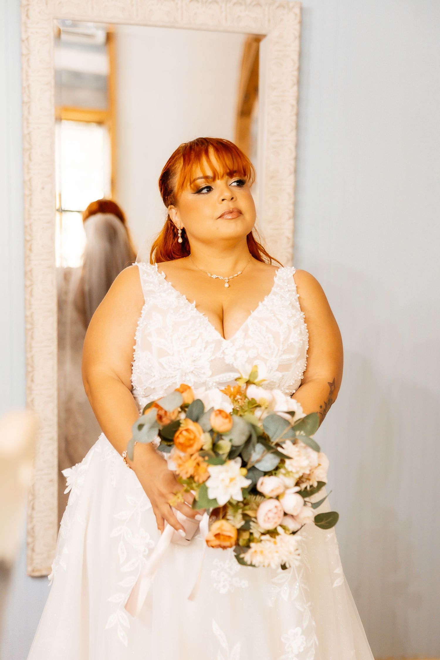 A bride in a white lace V-neck wedding gown holds a peach and white floral bouquet while posing in front of a mirror.