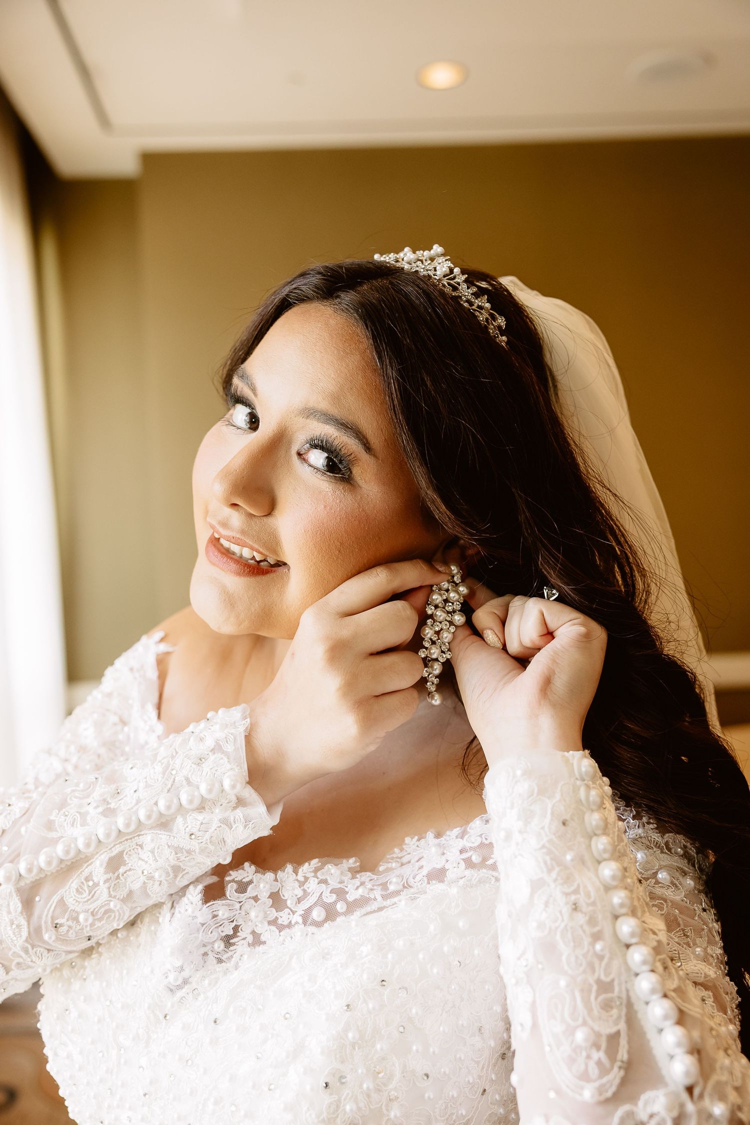 A bride in a long-sleeved white lace wedding dress with pearl details puts on her earrings before the ceremony.