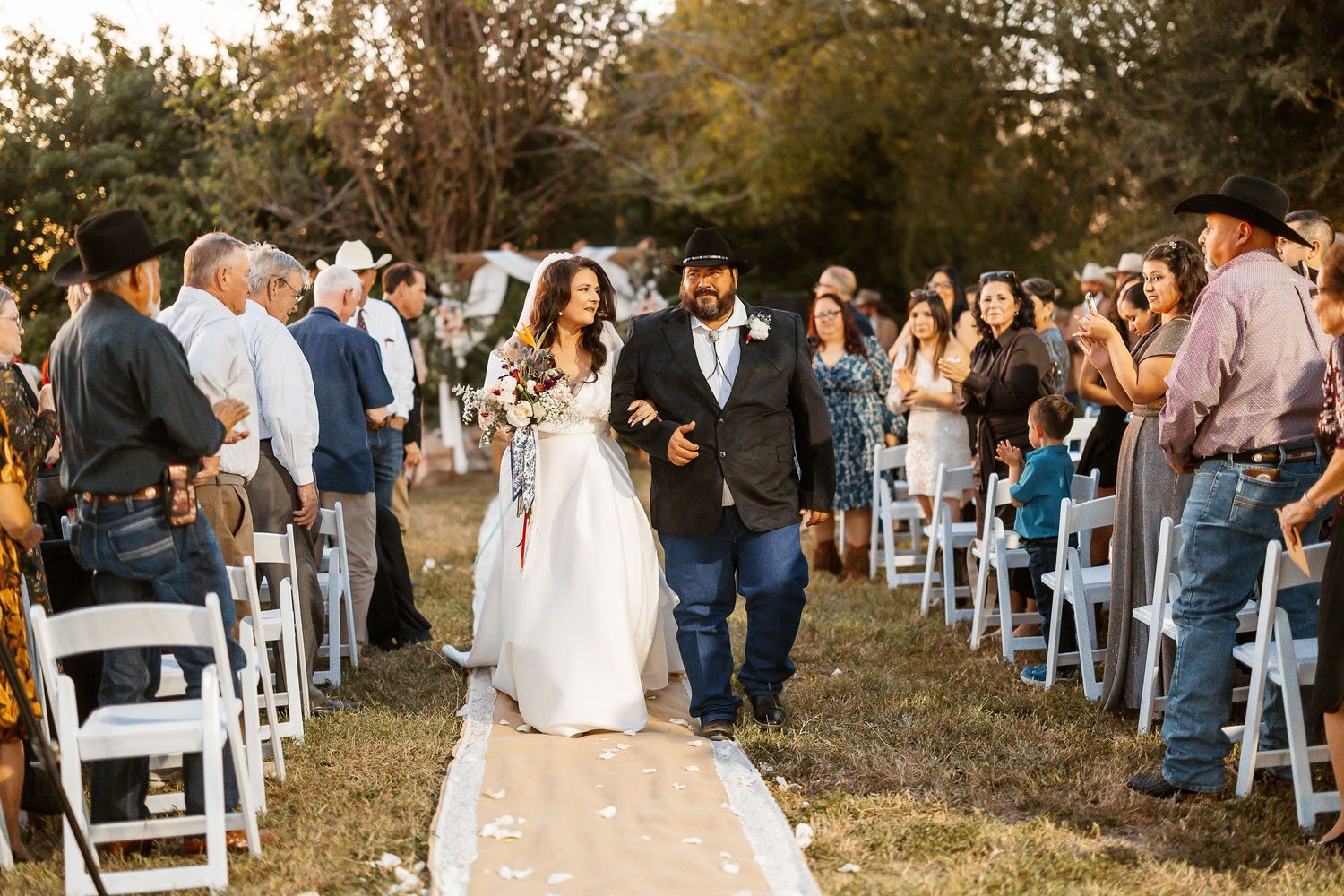 A beautiful outdoor wedding ceremony with guests seated in white chairs along a tree-lined path during golden hour.
