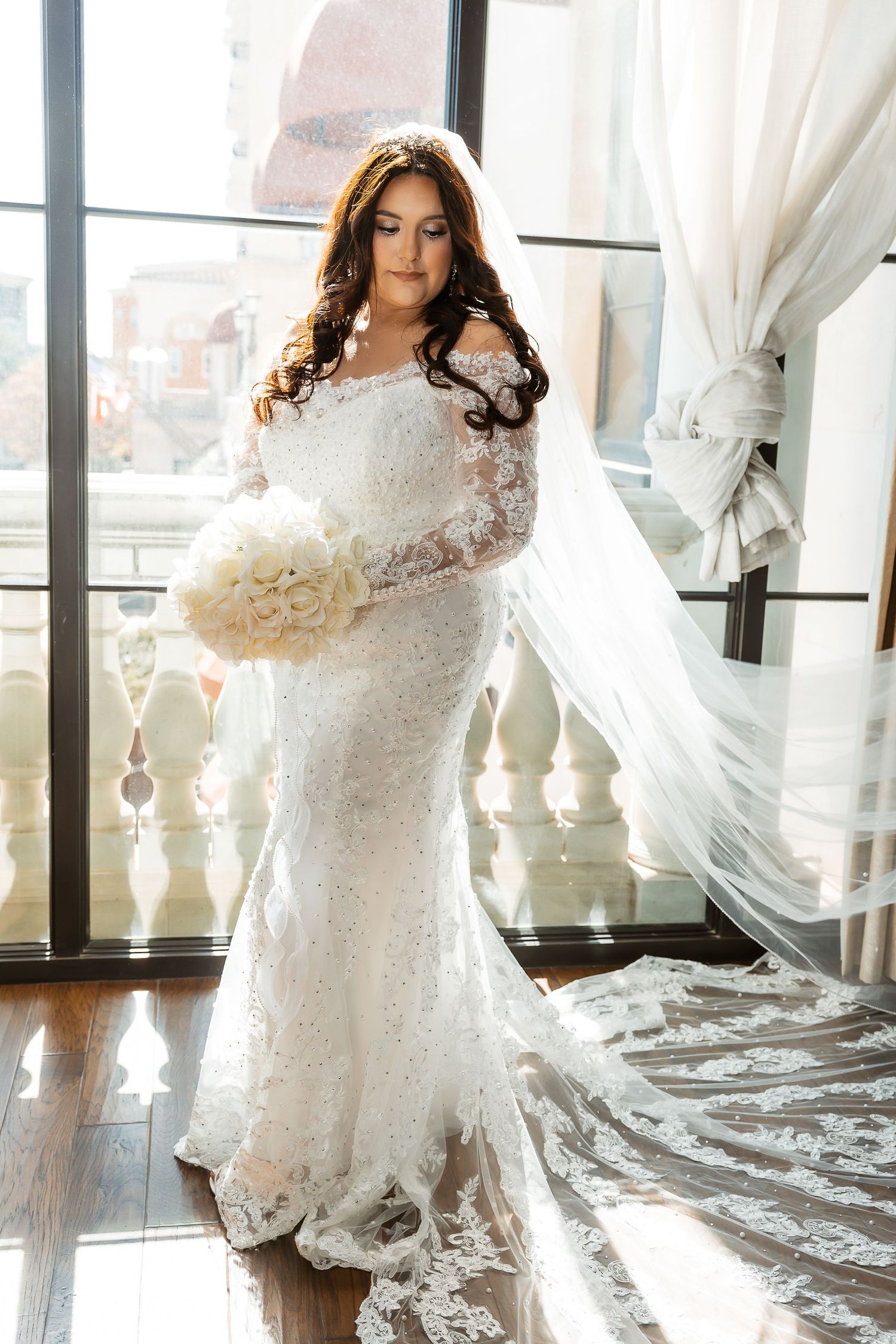 Bride in elegant lace wedding gown poses gracefully by sunlit windows with flowing white curtains in luxury venue.