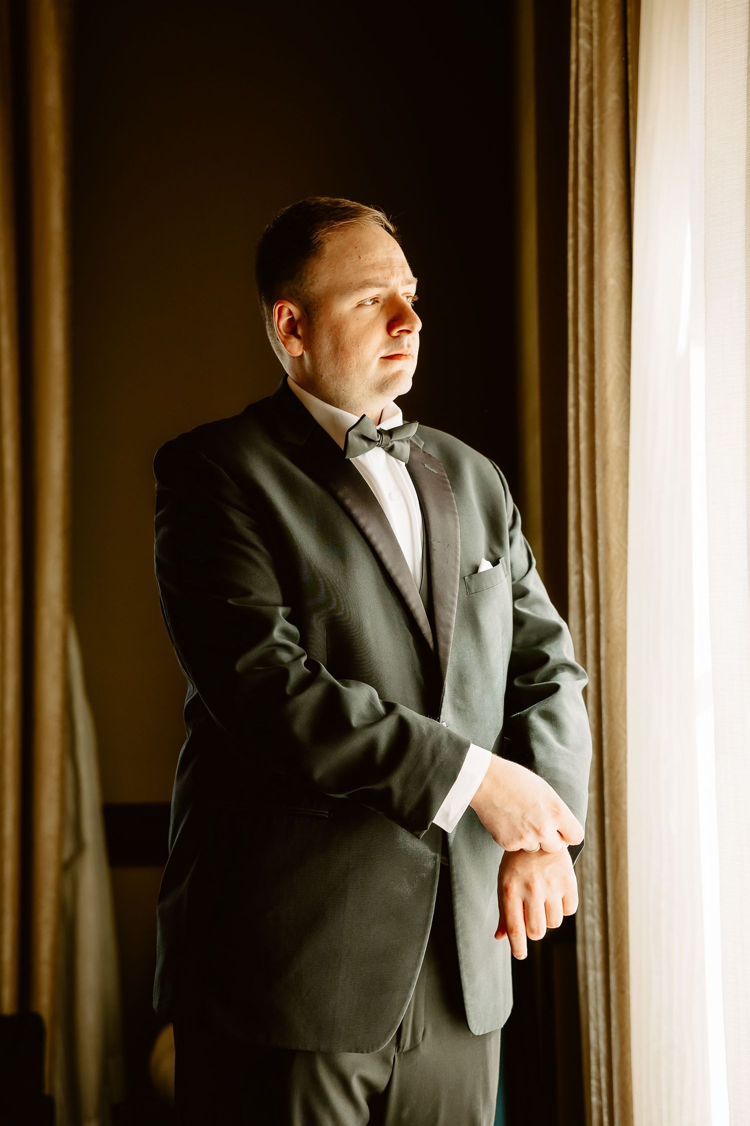 A person in a formal black tuxedo and bow tie stands contemplatively by a window in dramatic lighting.