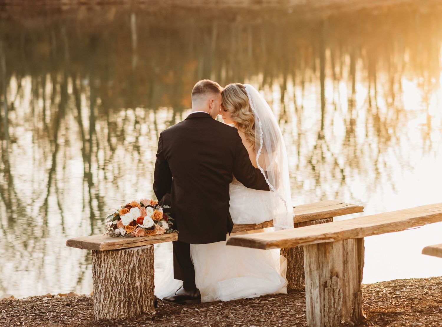 Bride and groom are sitting on a bench next to a lake with their backs to the camera and their foreheads touching