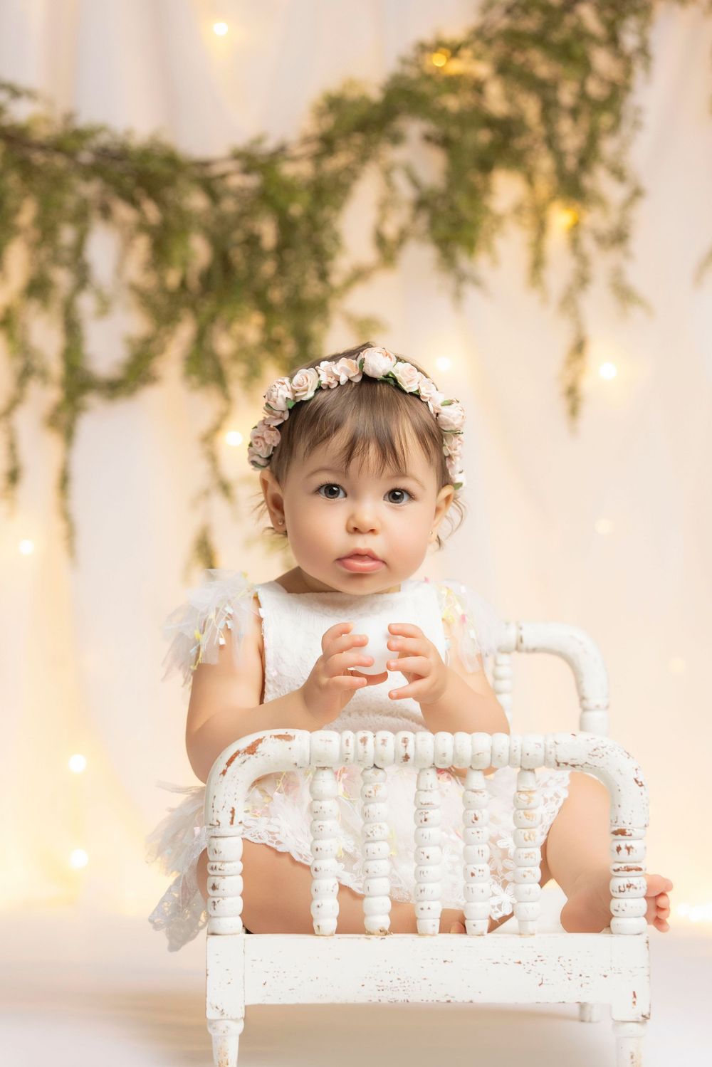 A young girl wearing a flower crown sits in a white chair, surrounded by greenery and twinkling lights.