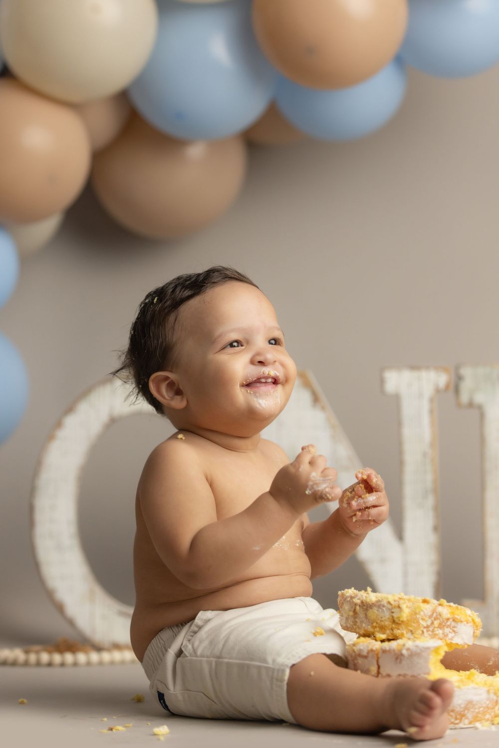 Happy baby smashing first birthday cake surrounded by blue and beige balloon decorations during cake smash photo session.