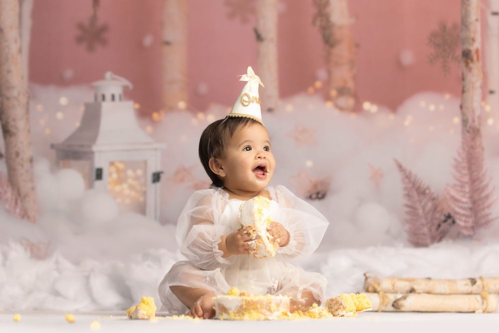 A baby wearing a party hat sits on a snowy scene with toys, surrounded by a winter landscape.