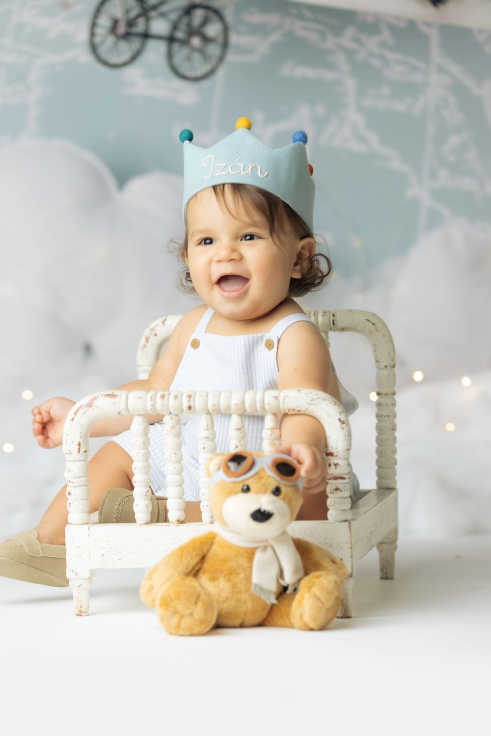 A happy baby wearing a crown and holding a stuffed animal, sitting in a white chair with a colorful background.