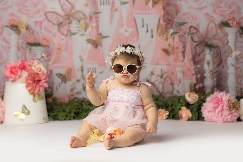 A baby girl wearing a pink dress and sunglasses sits among flowers and floral decor.