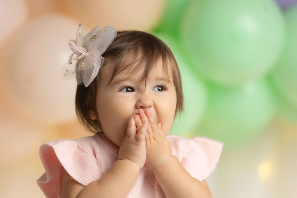 A young girl in a pink dress covers her mouth with her hands, with colorful balloons in the background.
