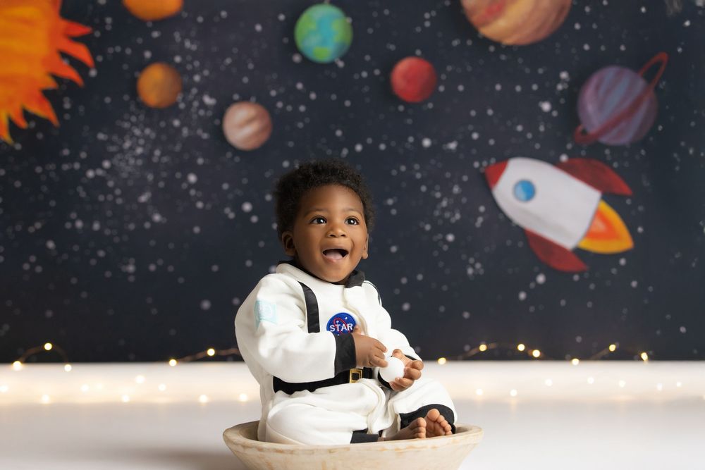 Happy baby in NASA astronaut outfit sits in wooden bowl before colorful space backdrop with planets and rocket.