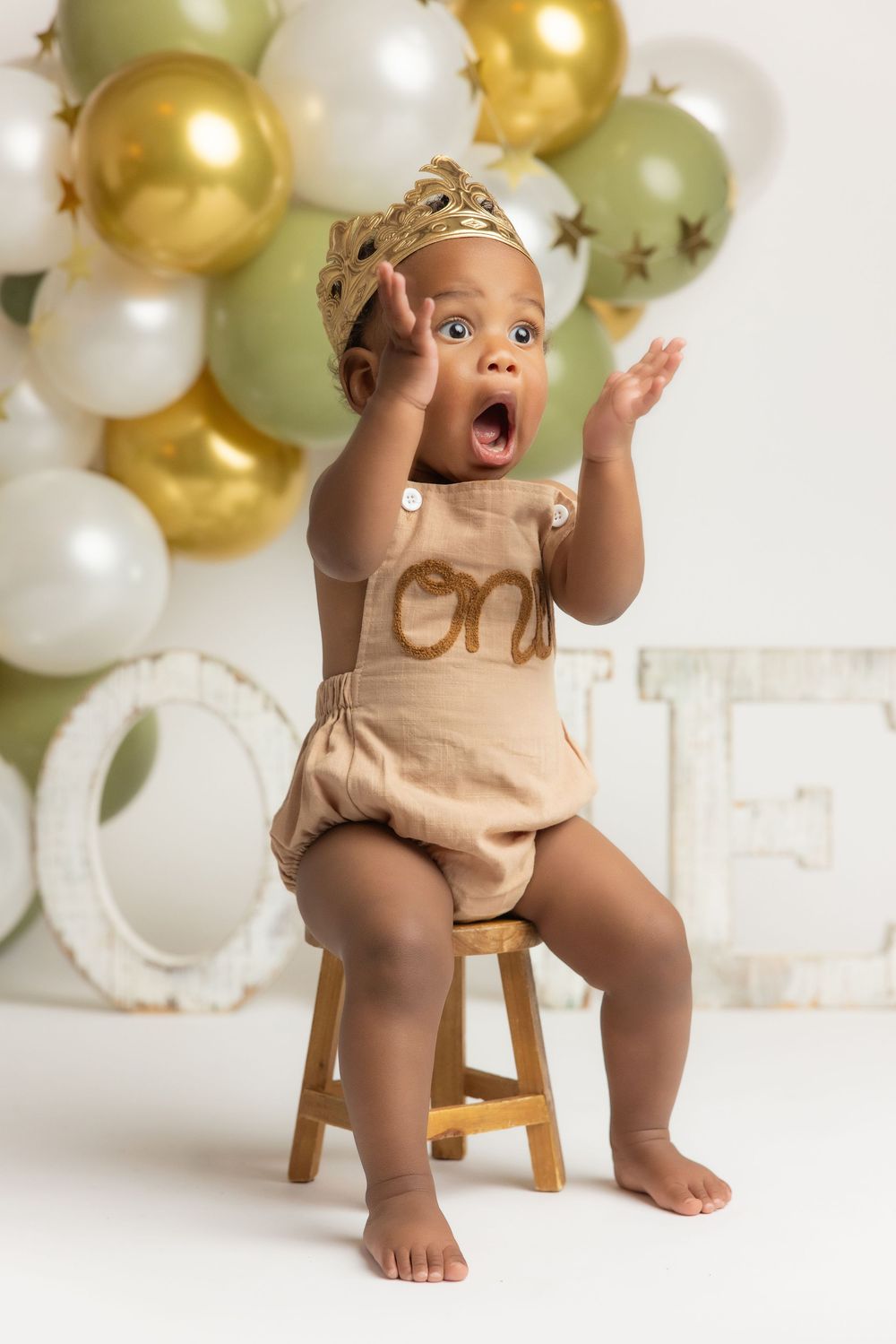 A baby in a beige outfit sitting on a stool and making a surprised facial expression surrounded by gold and white balloons.