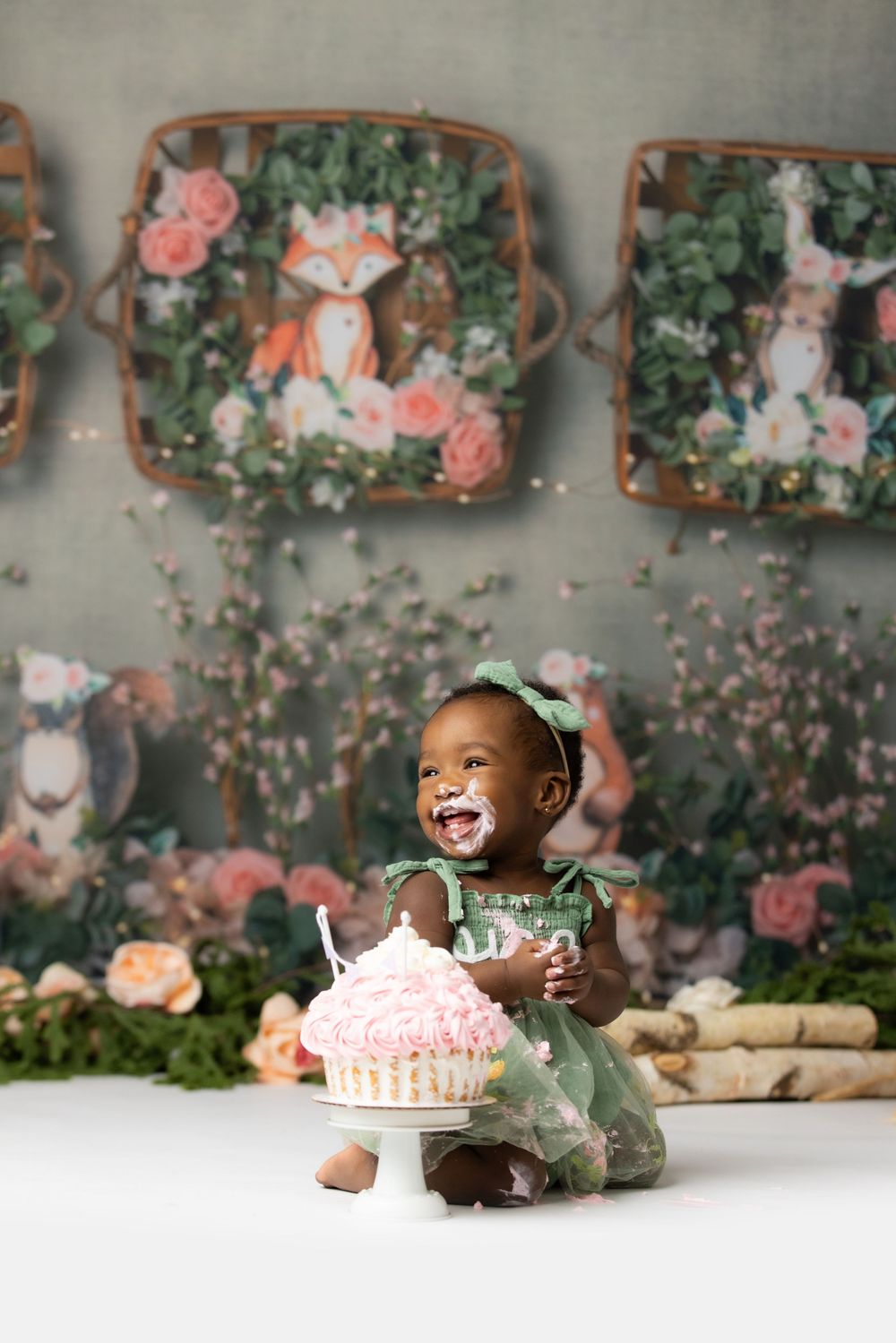 A smiling baby girl in a floral dress sits on a table in front of decorative floral artwork.