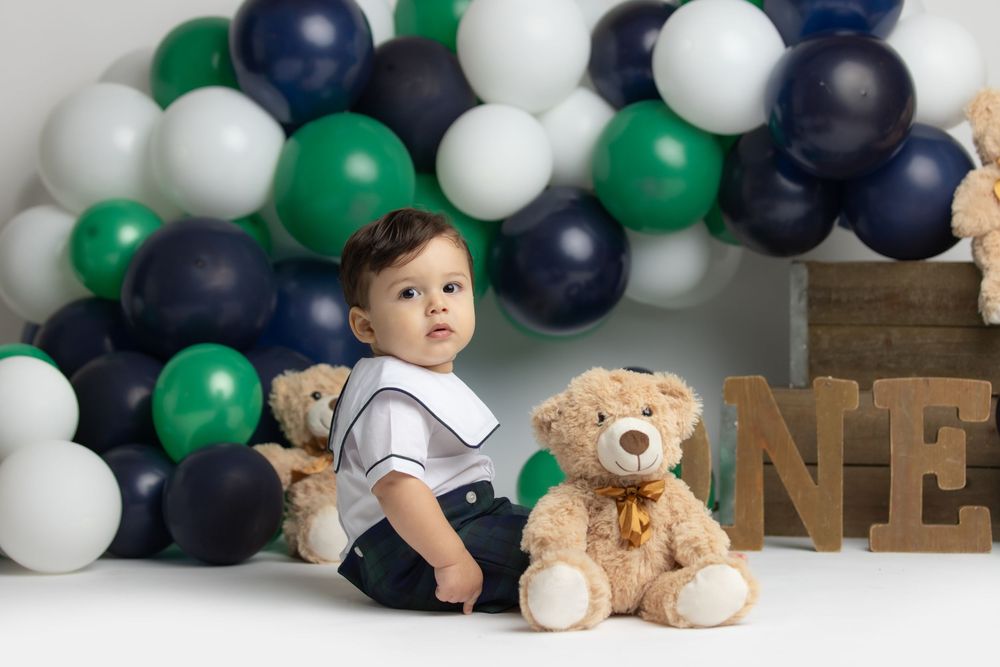 A young child sitting on the floor surrounded by colorful balloons and holding a stuffed animal.
