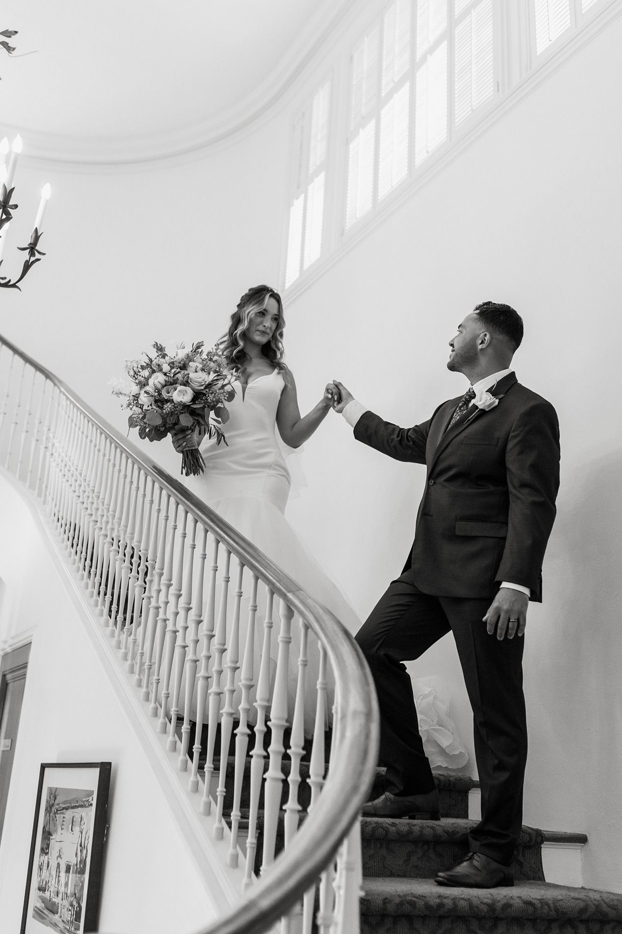 Black and white photo of a bride and groom holding hands on an elegant curved staircase, bride holding a bouquet at Darlington House.