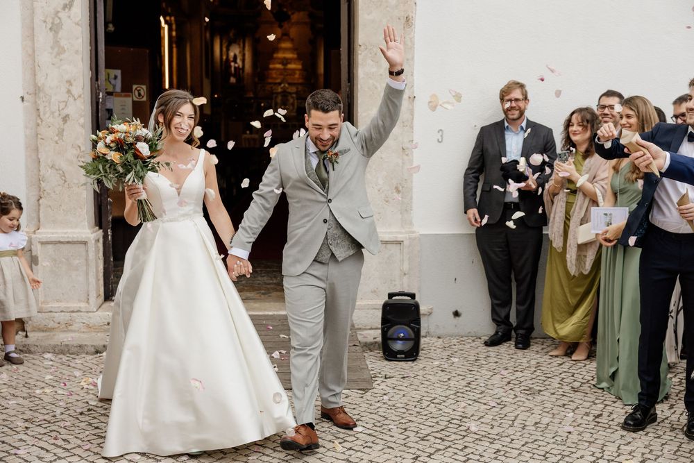 Newlywed couple exits church ceremony as guests toss flower petals, bride in white gown, groom in gray suit.